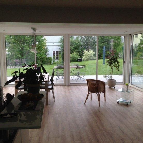 Ein offenes modernes Wohnzimmer mit Glaswänden und Türen, die zu einer Terrasse führen. Topfpflanzen, ein Esstisch und Stühle sind im Inneren, mit einem Mann, der sich neben den Glastüren bewegt.