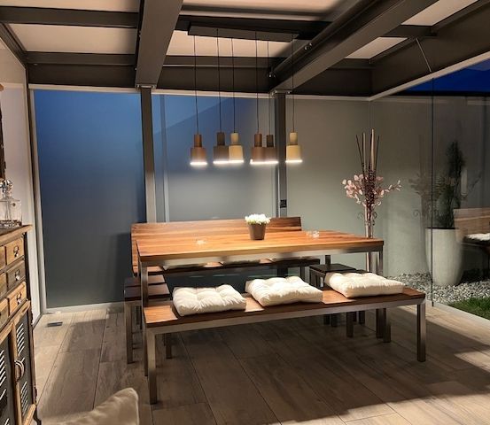 Ein Innenbereich zum Essen mit einem Holztisch und Bänken, geschmückt mit weißen Kissen, umgeben von einer blauen Wand und Glasfenstern.
