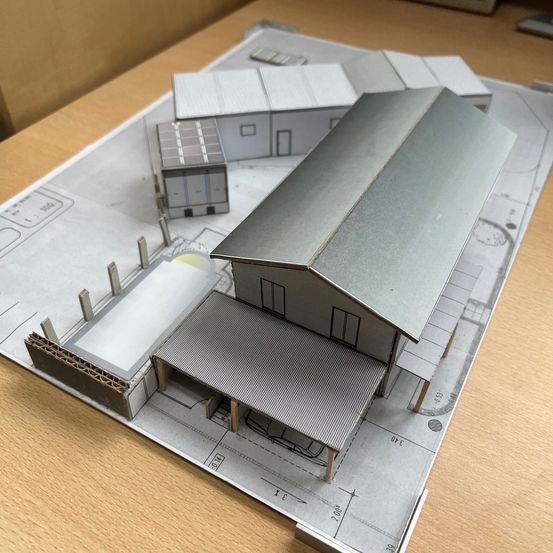 Ein Modell eines Gebäudes mit Carport steht auf einem Holztisch. Das Gebäude hat ein großes Metalldach und ist von anderen Strukturen umgeben.