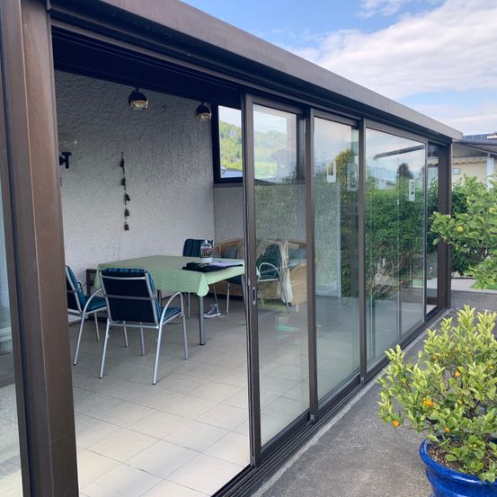Ein Patio mit gläsernen Schiebetüren und einem Esstisch mit Stühlen. Topfpflanzen stehen auf beiden Seiten des Patios. Der Patio hat einen gefliesten Boden.