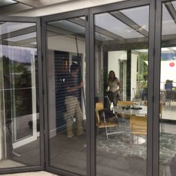 Ein Mann steht neben einer Schiebetür aus Glas, und eine Frau steht bei einem Tisch. Die Szene spielt sich in einem Haus mit einer Terrasse ab.