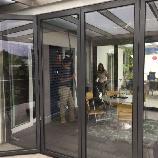 Ein Mann steht neben einer Schiebetür aus Glas, und eine Frau steht bei einem Tisch. Die Szene spielt sich in einem Haus mit einer Terrasse ab.