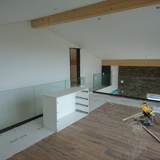 Ein leerer Raum mit Holzböden, weißen Wänden und einer Glasbrüstung. Ein weißer Schrank mit Schubladen steht in der Nähe der Wand. Ein gelbes Elektrowerkzeug liegt auf dem Boden.