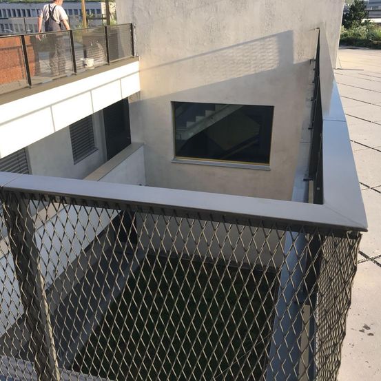 Ein Gebäude mit Balkon hat ein Fenster an der Wand, und eine Person steht darauf. Das Gebäude hat eine Treppe und einen Zaun.