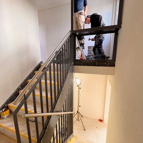 Zwei Arbeiter in einem Treppenhaus, einer steht auf einer Leiter und repariert das Geländer. Eine Lampe steht auf dem Boden.