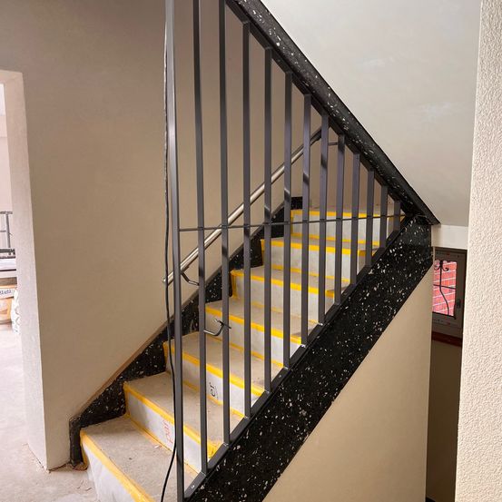 Eine Treppe mit schwarz-gelbem Stufenmuster, schwarzen Geländern und einer teilweise abgedeckten Wand. Die Wand und die Decke scheinen unfertig zu sein.
