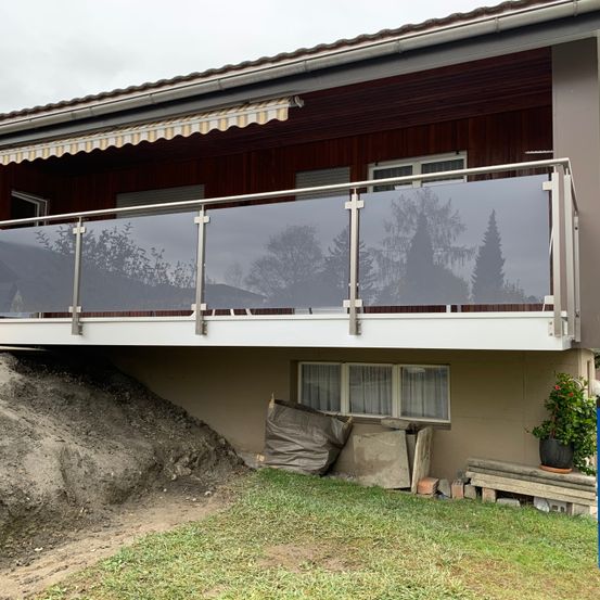 Ein Haus mit Balkon hat ein Glasgeländer. Der Balkon hat ein weißes Markisentuch. Unter dem Balkon befinden sich Schmutzhaufen, ein Topf mit Pflanzen und einige Holzbretter.