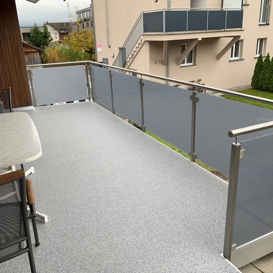 Ein Balkon mit grauem Boden und Metallgeländern, ausgestattet mit einem Tisch und Stühlen. Der Balkon befindet sich im oberen Stockwerk eines Gebäudes mit einer Treppe.