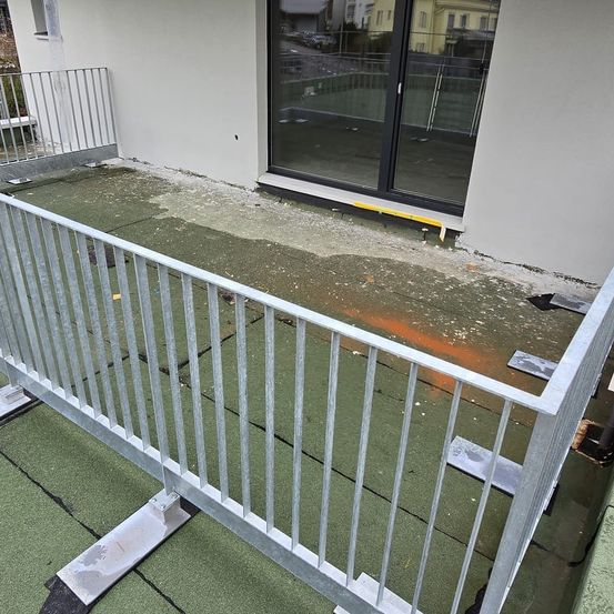 Ein Balkon mit einem Metallgeländer und gefliestem Boden. Neben dem Geländer befindet sich eine Glastür mit Reflexionen von Bäumen und einem Gebäude.