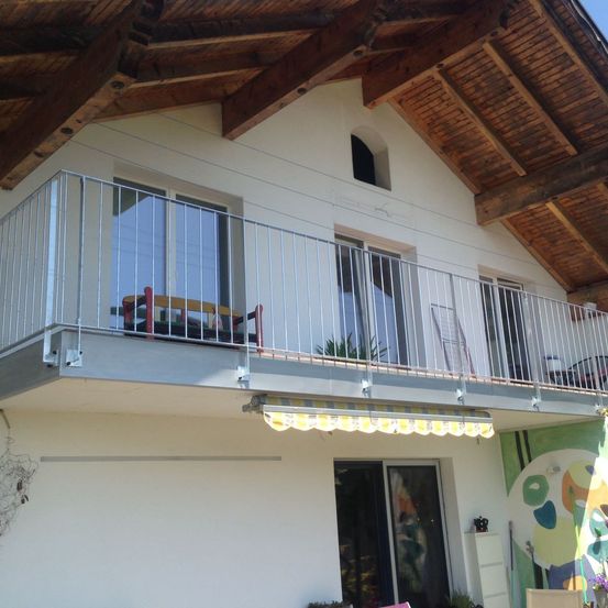 Ein Balkon eines weißen Hauses mit einem Metallgeländer und einem gelben Vordach. Ein Stuhl steht auf dem Balkon. Das Haus hat Glasfenster.