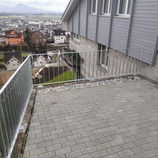 Ein Balkon mit Metallgeländer und Ziegelboden, mit Blick auf eine Stadt mit verschiedenen Gebäuden und Bergen in der Ferne.