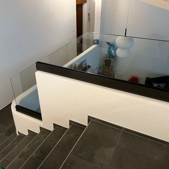 Eine moderne Treppe mit einer Glasbrüstung, schwarzem Rand und weißen Wänden. Sie hat mehrere Stufen, die nach oben führen. Darüber befindet sich eine Glasdecke mit einer hängenden Lampe.