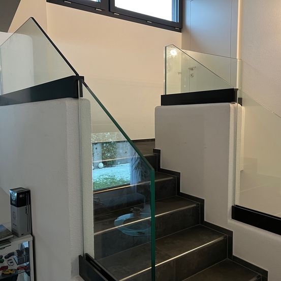 Eine moderne Treppe mit Glasstufen und einem Glasgeländer. Die Treppe hat ein schlankes, minimalistisches Design mit weißen Wänden und schwarzen Akzenten. Das Glasgeländer bietet einen klaren Blick auf den Außenbereich.