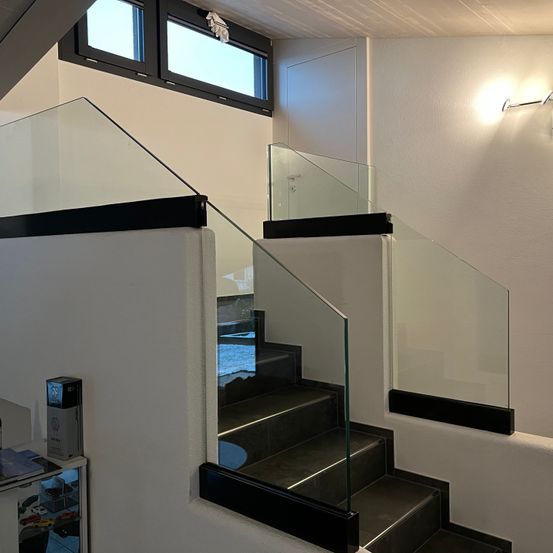 Eine Innentreppe mit Glasstufen und einem schwarzen Geländer. Die Wände sind weiß und es gibt ein Glasgeländer entlang der Stufen.