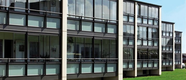 Ein mehrstöckiges Gebäude mit vielen Glasfenstern und schwarzen Rahmen, auf einem Rasen mit einem klaren blauen Himmel im Hintergrund gelegen.