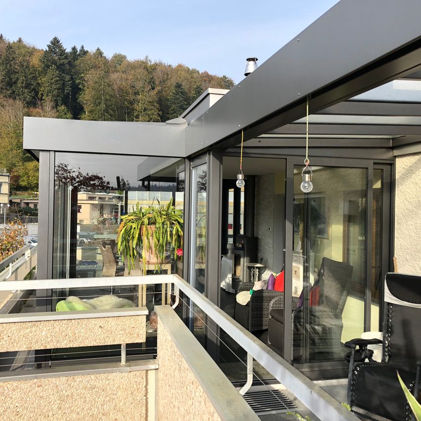 Ein Balkon mit einer Glaswand, einem Metallgeländer und hängenden Lampen. Im Inneren befinden sich ein Stuhl, eine Couch und eine Topfpflanze. Bäume und Gebäude sind im Hintergrund zu sehen.