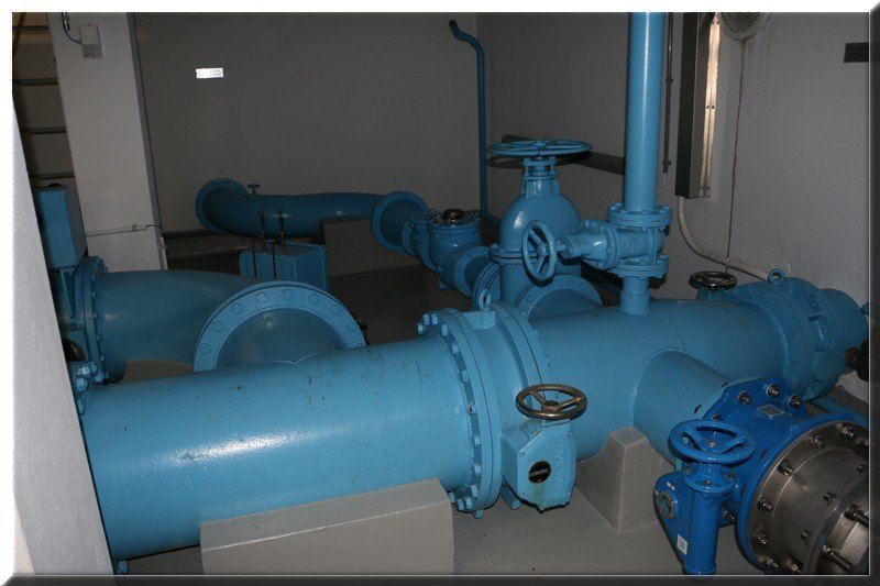 A room with multiple blue pipes and valves, some of which have handles. The pipes are connected to each other and to the wall.