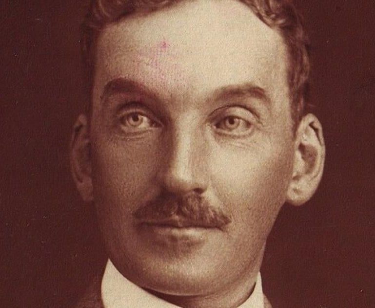 An old sepia-toned portrait shows a man with a mustache and short, curly hair. He wears a white collar and appears to be looking directly at the camera.
