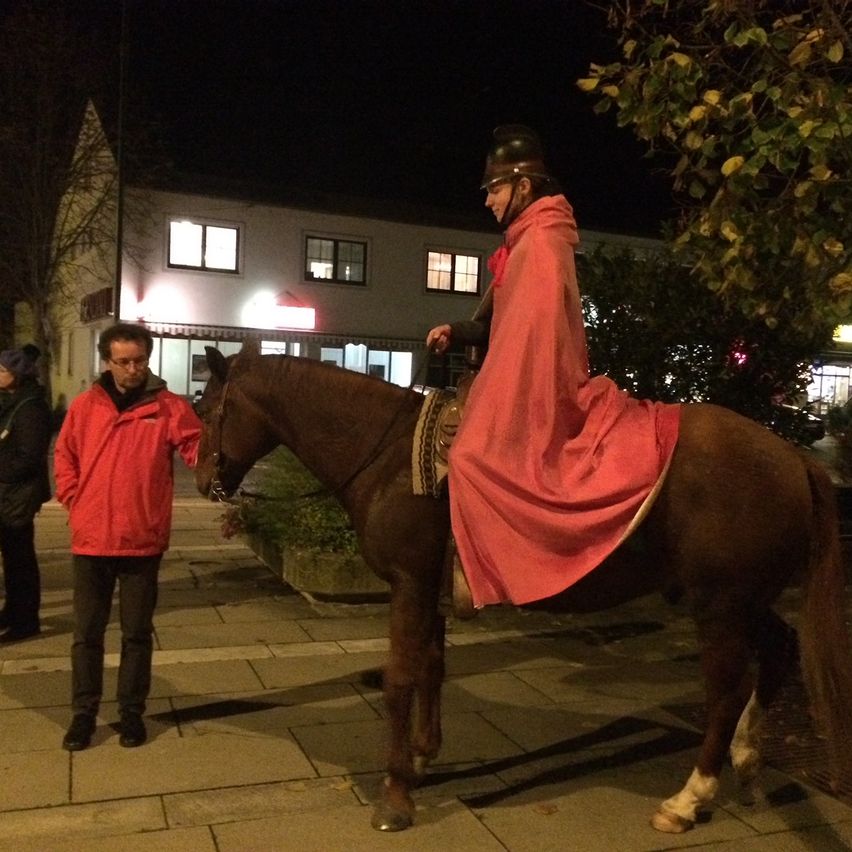 A man dressed in medieval attire rides a horse in the evening, with another man in a red jacket observing.