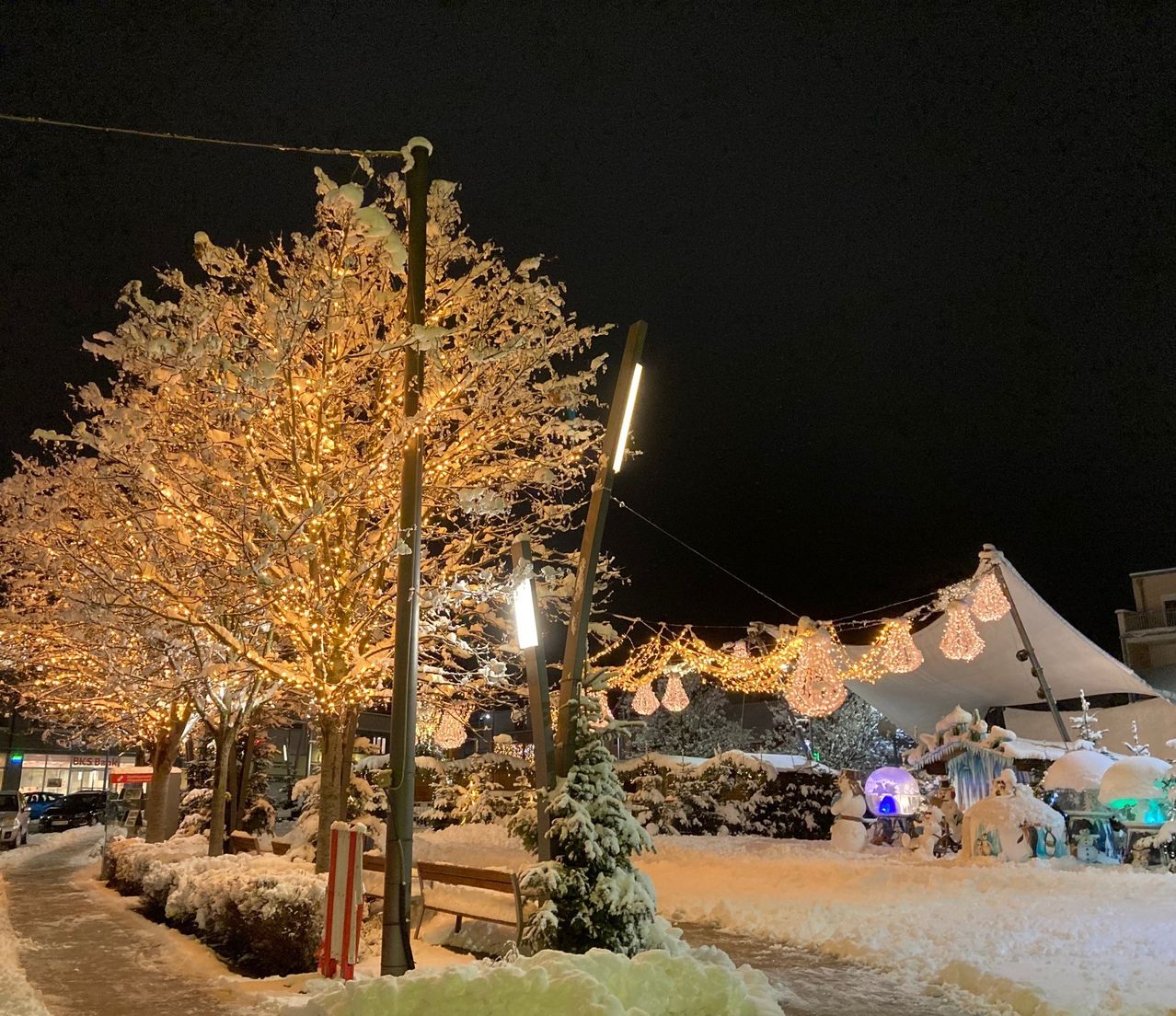 Eine verschneite Straße mit Bäumen, die mit Lichtern und Ornamenten geschmückt sind. Ein festlicher Zeltaufbau mit Lichtern und Ornamenten im Hintergrund. Ein Schneemann und andere Winterdekorationen sind vorhanden.
