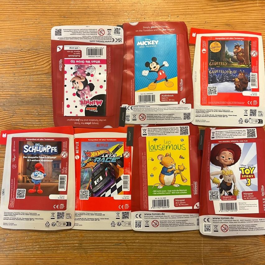 Eine Sammlung roter Plastiktüten mit verschiedenen Kinderfilm- und Buchcovern auf einem Holztisch. Tüten beinhalten Minnie Maus, Micky Maus, Gruffelo, Schlumpf, Hot Wheels, Leo der Maus und Toy Story.