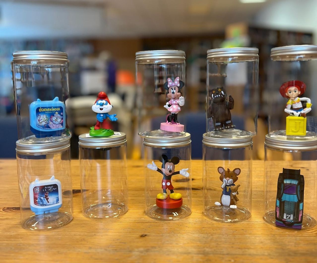 Sechs durchsichtige Gläser mit Deckeln stehen auf einem Holztisch. In jedem Glas befinden sich Figuren von Micky Maus, Minnie Maus und anderen Charakteren.