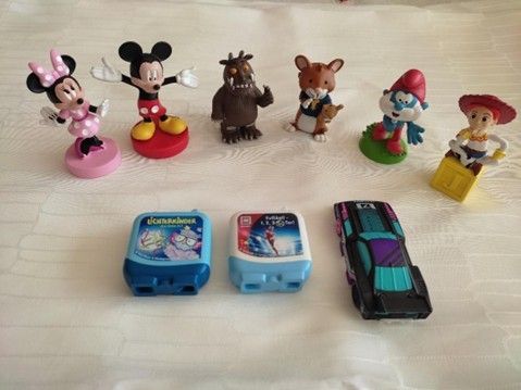 Eine Sammlung von Spielzeugfiguren und Fahrzeugen auf einer weißen Oberfläche: Minnie Maus, Micky Maus, eine braune Kreatur, eine braune Maus, ein Schlumpf, ein Cowboy, ein blau-weißes Spielzeug und ein schwarzes Spielzeugauto.