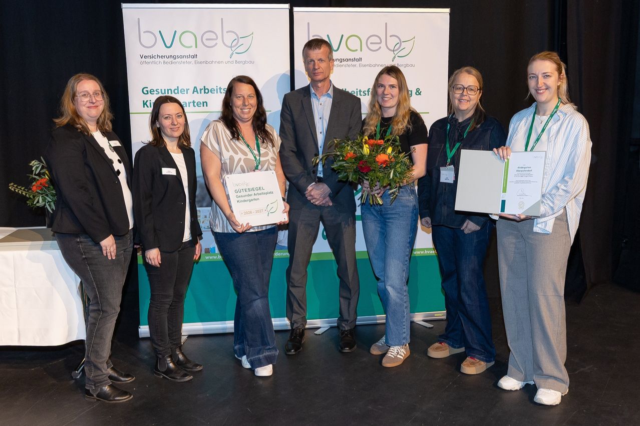 Eine Gruppe von Frauen und ein Mann stehen vor einem Banner. Sie halten Auszeichnungen und einen Blumenstrauß. Sie alle lächeln.