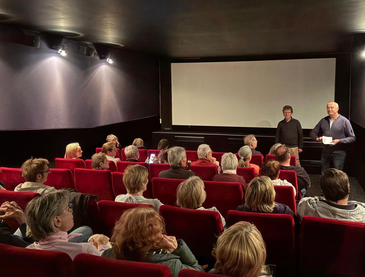 Eine Gruppe von Menschen sitzt in roten Stühlen in einem Kino. Ein Mann steht vor einer Projektionsleinwand, während ein anderer Mann hinter ihm steht.