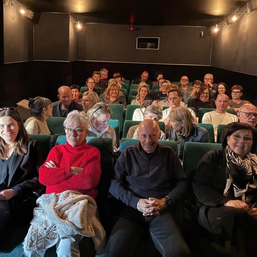 Eine Gruppe von Menschen mit Brillen und einigen mit Schals sitzt im Auditorium. Der Mann vorne lächelt und hat die Hände gefaltet.