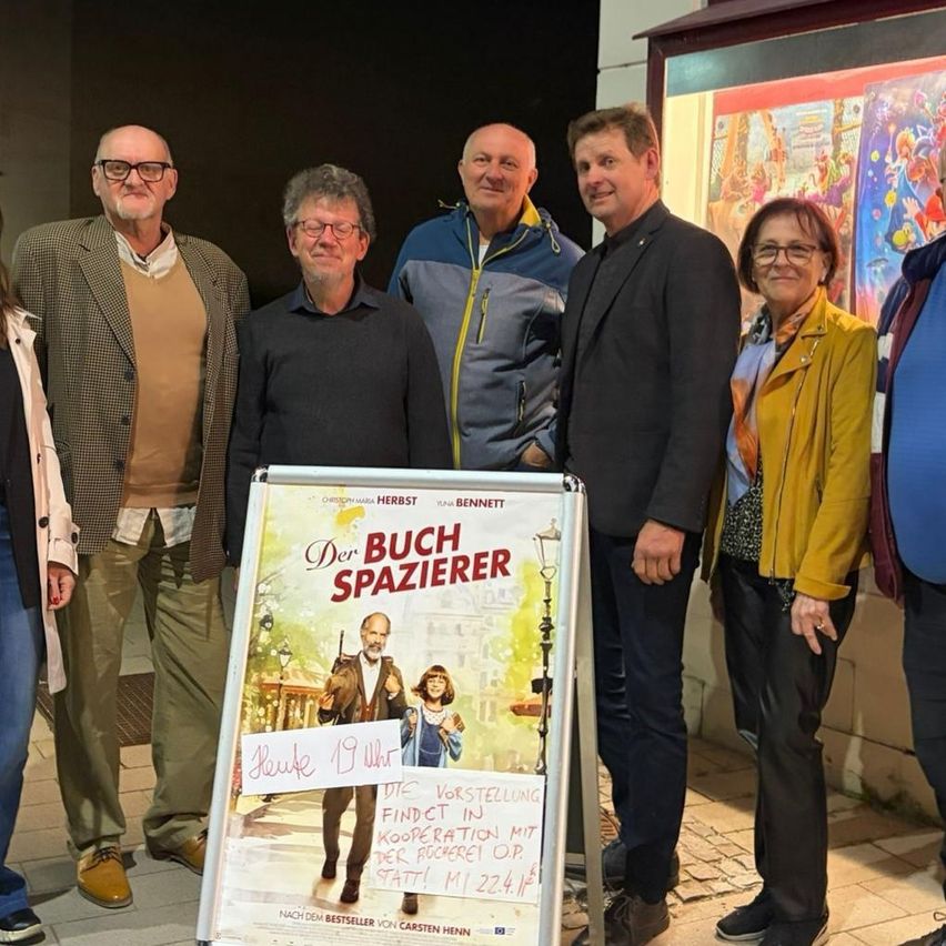 Eine Gruppe von Menschen steht zusammen vor einem Plakat, das 'Der Buchspazierer' bewirbt. Sie lächeln und scheinen für ein Foto zu posieren.