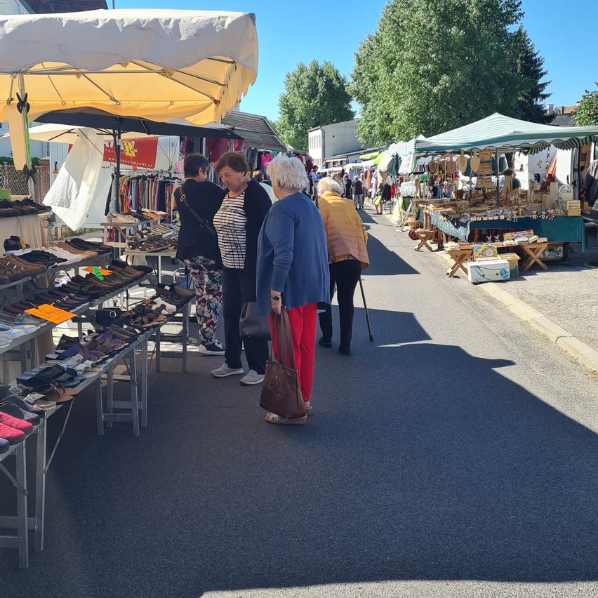 Eine Gruppe von Menschen steht unter einem Regenschirm auf einem Straßenmarkt und betrachtet verschiedene Schuhe in der Ausstellung. Bäume und Stände säumen die Straße, ein Gebäude ist in der Ferne sichtbar.