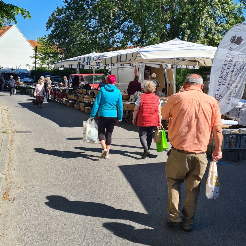 Ein Straßenmarkt an einem sonnigen Tag. Leute kaufen unter Schirmen, mit Ständen, die verschiedene Artikel verkaufen. Ein Mann und eine Frau gehen am Markt vorbei.