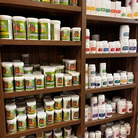 Ein hölzerner Regal mit verschiedenen Flaschen und Behältern zur Ausstellung. Die linke Seite hat einen Stapel weisser und grüner Behälter mit der Aufschrift 'lebe natur' und Schmetterlingsbildern. Die rechte Seite hat mehrere Reihen von weissen und unterschiedlich farbigen Flaschen mit Etiketten wie 'Magnesium Citrat' und 'K2 Vitamin'.