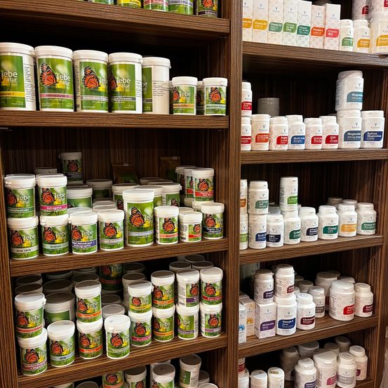 Ein hölzerner Regal mit verschiedenen Flaschen und Behältern zur Ausstellung. Die linke Seite hat einen Stapel weisser und grüner Behälter mit der Aufschrift 'lebe natur' und Schmetterlingsbildern. Die rechte Seite hat mehrere Reihen von weissen und unterschiedlich farbigen Flaschen mit Etiketten wie 'Magnesium Citrat' und 'K2 Vitamin'.