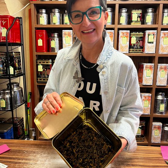 Eine Frau in einer weißen Jeansjacke und mit Brille hält eine Blechdose mit losen Teeblättern in einem Geschäft mit Regalen, die mit Teedosen gefüllt sind.