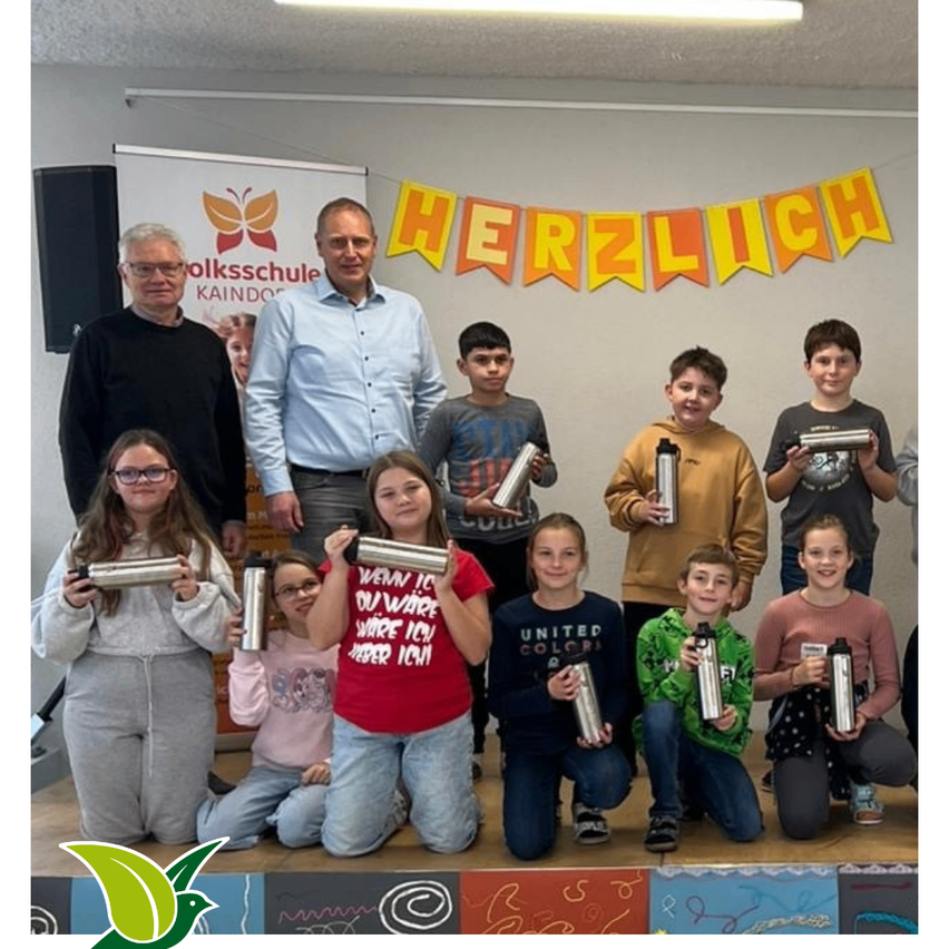 Eine Gruppe von Schülern und Lehrern steht mit silbernen Flaschen auf der Bühne, einem weißen Banner mit Schmetterlingslogo und einem gelben Banner, das Herzlich sagt.