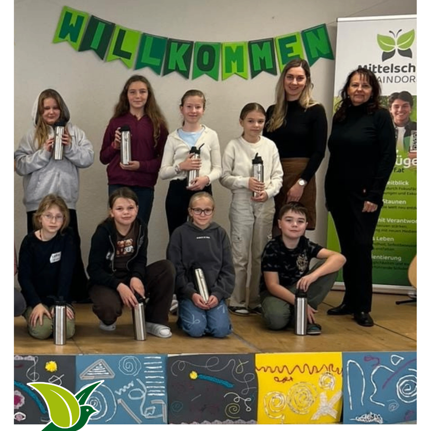 Eine Gruppe von Kindern und Erwachsenen posiert für ein Foto mit Edelstahlbechern. Sie stehen auf einer Bühne mit farbenfrohen Kunstwerken im Hintergrund.