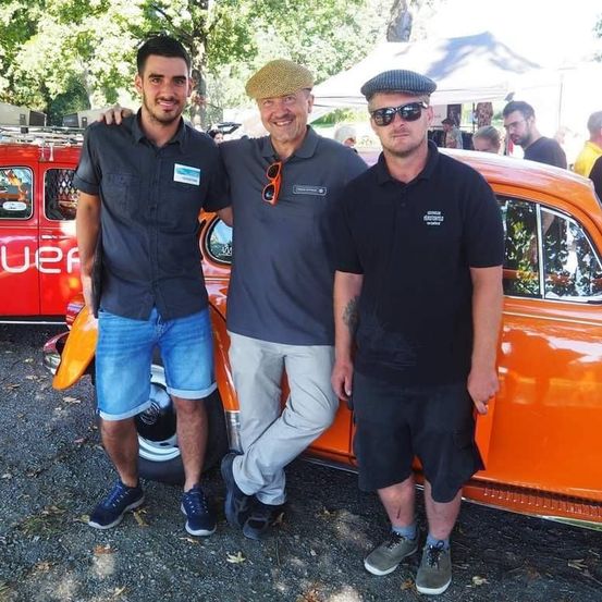 Three men stand beside an orange vintage car with a luggage rack on the roof, wearing casual attire. They smile for a photo in an outdoor setting with trees and tents in the background.