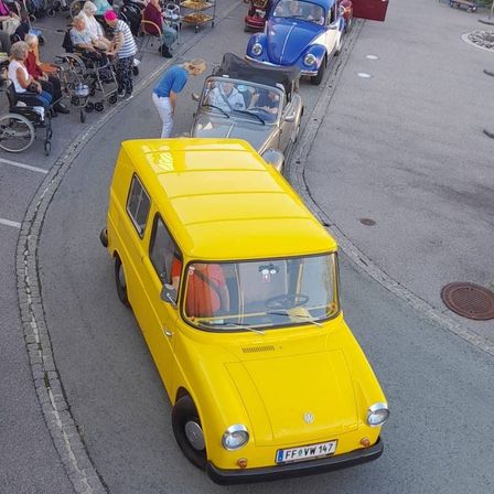 Ein gelbes Auto fährt auf einer Straße, an der ein blaues Auto und ein silbernes Auto geparkt sind. Mehrere Menschen in Rollstühlen und andere sitzen entlang der Seite.