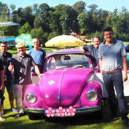 Fünf Männer posieren mit einem rosa Oldtimer, drei stehen davor und zwei lehnen sich an das Auto. Sie befinden sich in einem Rasenbereich mit Bäumen im Hintergrund.