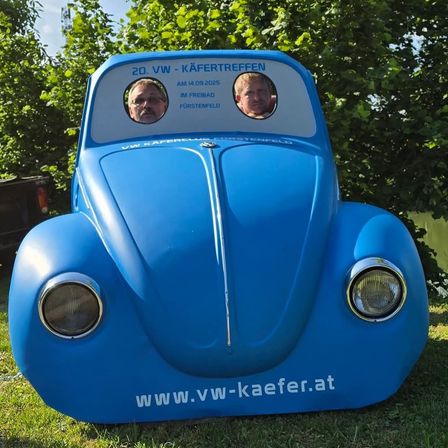 Ein blauer VW Käfer mit zwei Personen im Inneren, auf Gras geparkt. Das Auto ist mit dem Text '20. VW - Käfertreffen' verziert.