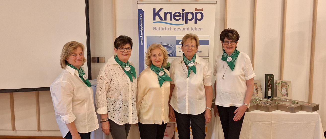 Vier Frauen mit grünen Schals stehen vor einem Banner, das 'Kneipp. Natürlich gesund leben.' besagt.