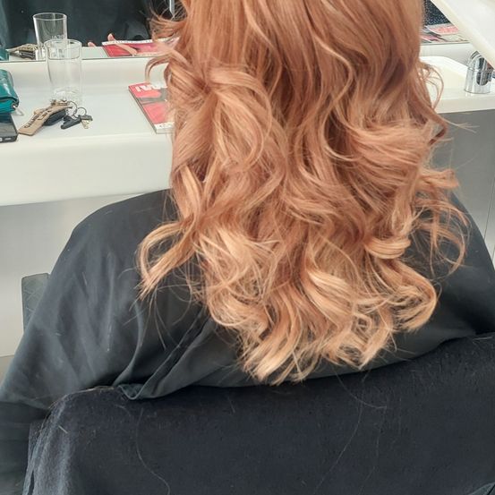 Eine Frau mit langen blonden Haaren sitzt in einem Salonstuhl. Sie schaut in einen Spiegel, hinter dem sich ein Schreibtisch befindet, auf dem Schlüssel, ein Telefon und eine Zeitschrift liegen.