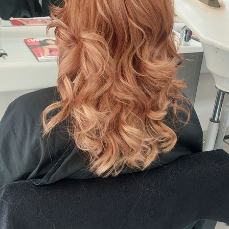 Eine Frau mit kupferfarbenen, gewellten Haaren sitzt in einem Salonstuhl und trägt einen schwarzen Umhang. Hinter ihr steht ein Schreibtisch mit einer Zeitschrift und Schlüsseln.