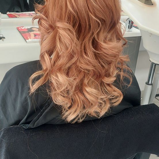 Eine Frau mit kupferfarbenen, gewellten Haaren sitzt in einem Salonstuhl und trägt einen schwarzen Umhang. Hinter ihr steht ein Schreibtisch mit einer Zeitschrift und Schlüsseln.