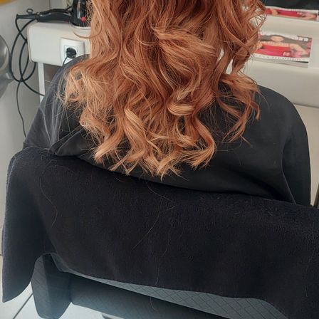 Eine Person mit lockigem orangefarbenem Haar sitzt in einem Stuhl in einem Salon. Die Person trägt ein schwarzes Oberteil. Dahinter befindet sich ein weißer Regalboden mit einer Zeitschrift darauf.