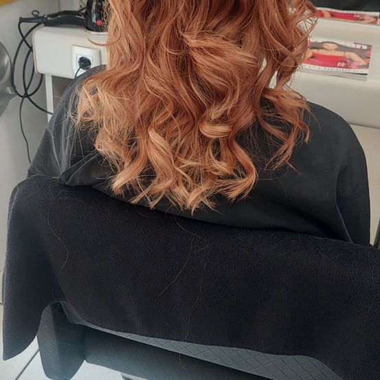 Eine Person mit lockigem orangefarbenem Haar sitzt in einem Stuhl in einem Salon. Die Person trägt ein schwarzes Oberteil. Dahinter befindet sich ein weißer Regalboden mit einer Zeitschrift darauf.