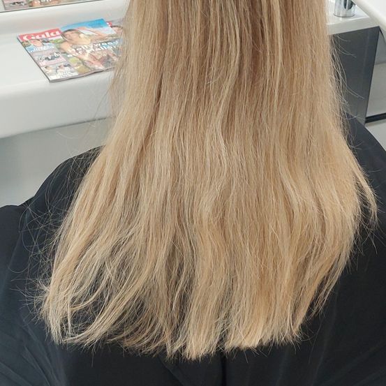 Eine Person mit langen blonden Haaren sitzt in einem Salon. Dahinter steht ein weißer Schreibtisch mit einer Zeitschrift namens 'Gula'.