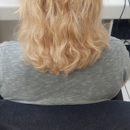 Eine Person mit langen blonden, gewellten Haaren sitzt in einem Stuhl und trägt ein graues T-Shirt, mit weißem Hintergrund.